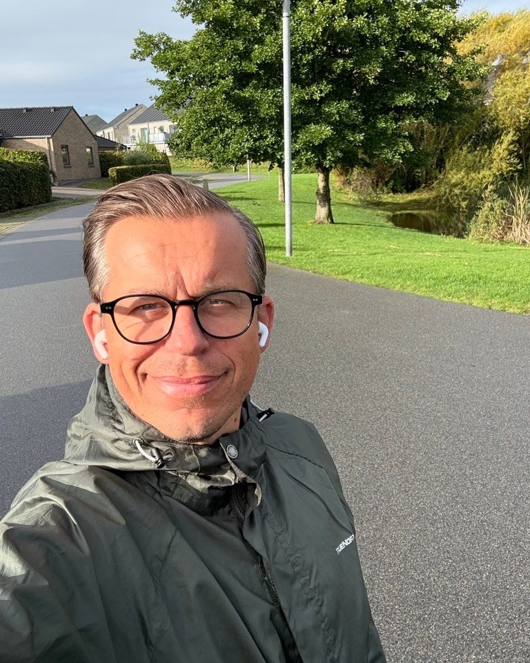 Lars on a walk, smiling into the camera with earbuds in, on a path between houses, a lawn and a pond.